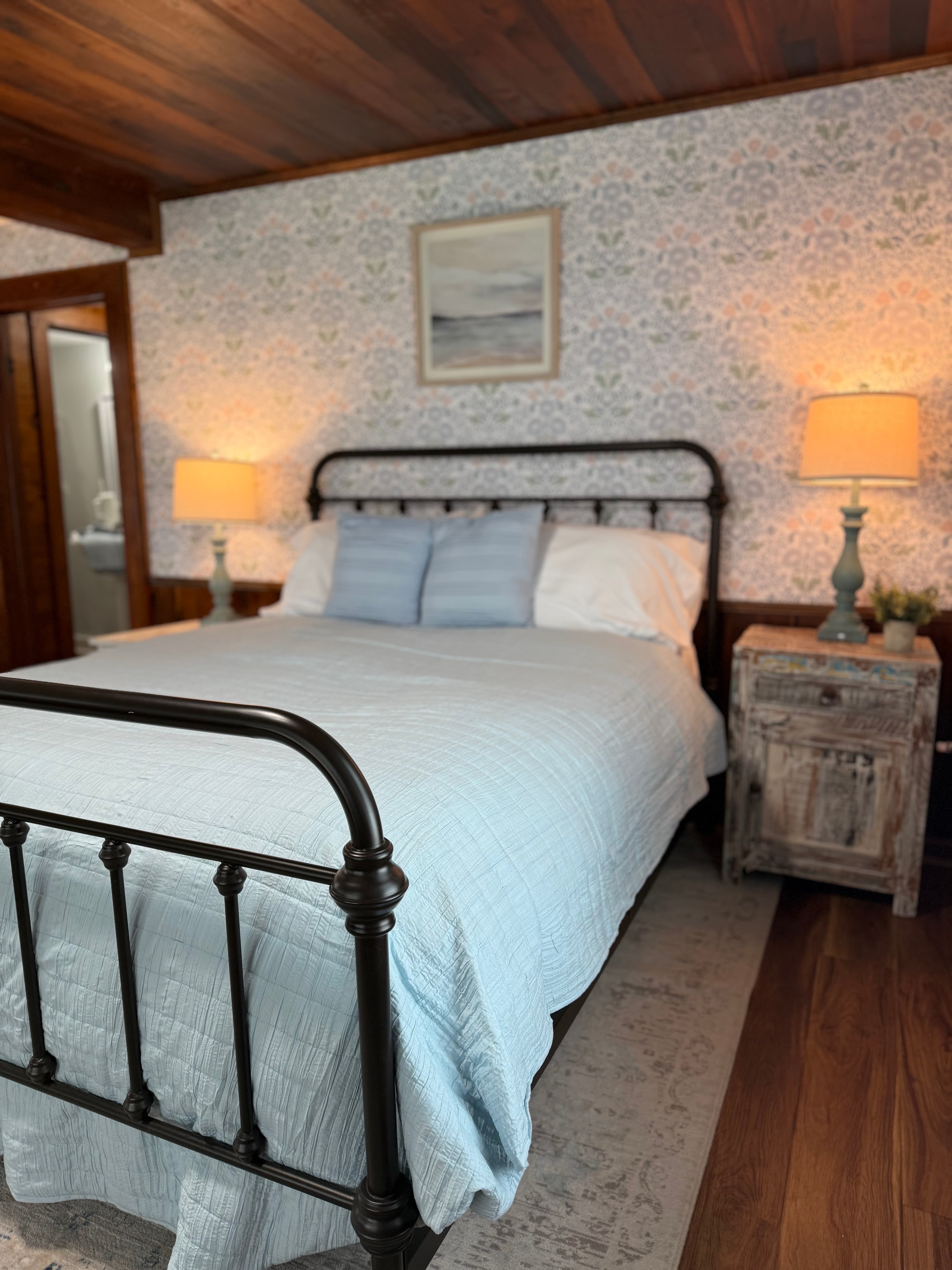 A detail view of a bedroom in a newly remodeled suite featuring a queen-size bed with an iron frame and light blue bedding. The room is decorated with coastal-patterned wallpaper, a rustic wooden nightstand with a lamp, and original dark wood ceilings and flooring.