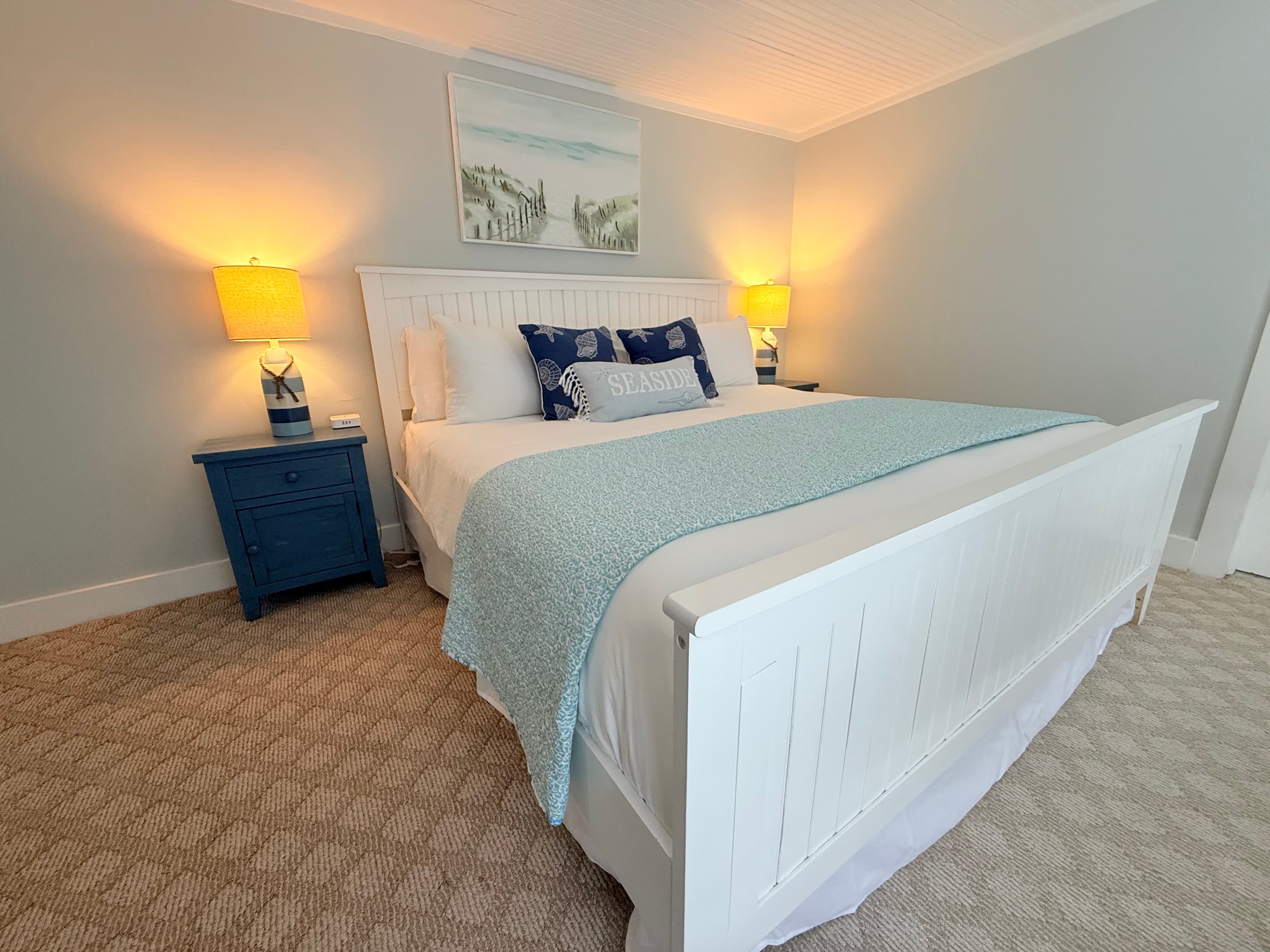 A guest room featuring a large white wooden bed frame with a navy and white striped comforter. The room includes a blue bedside table with a lamp, light gray walls, and a coastal landscape painting hanging above the headboard.