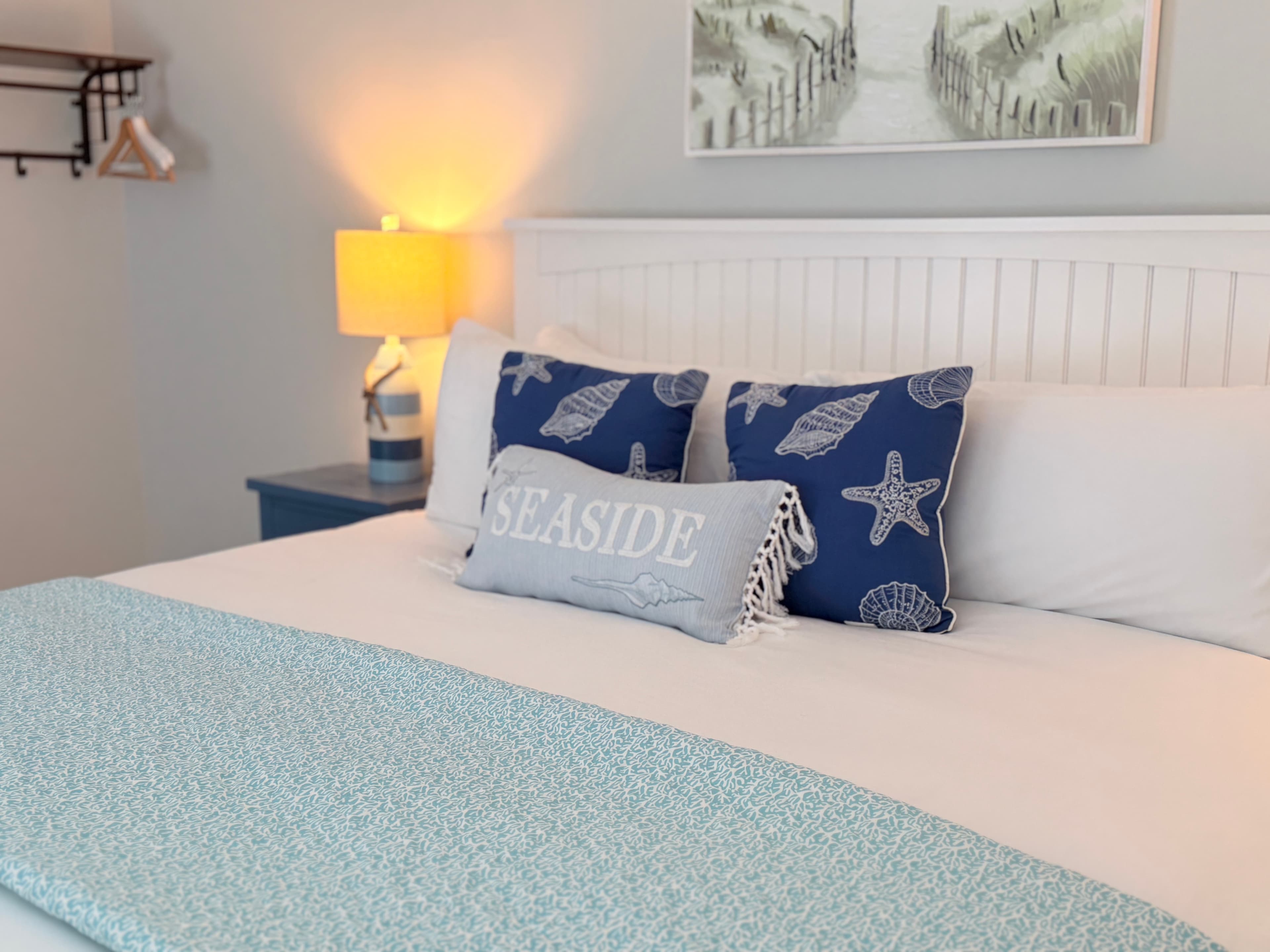 A brightly lit bedroom featuring a large bed with a white wooden headboard and a navy and white striped comforter. The room includes blue starfish accent pillows, light gray walls, and a coastal painting of sand dunes hanging above the bed.