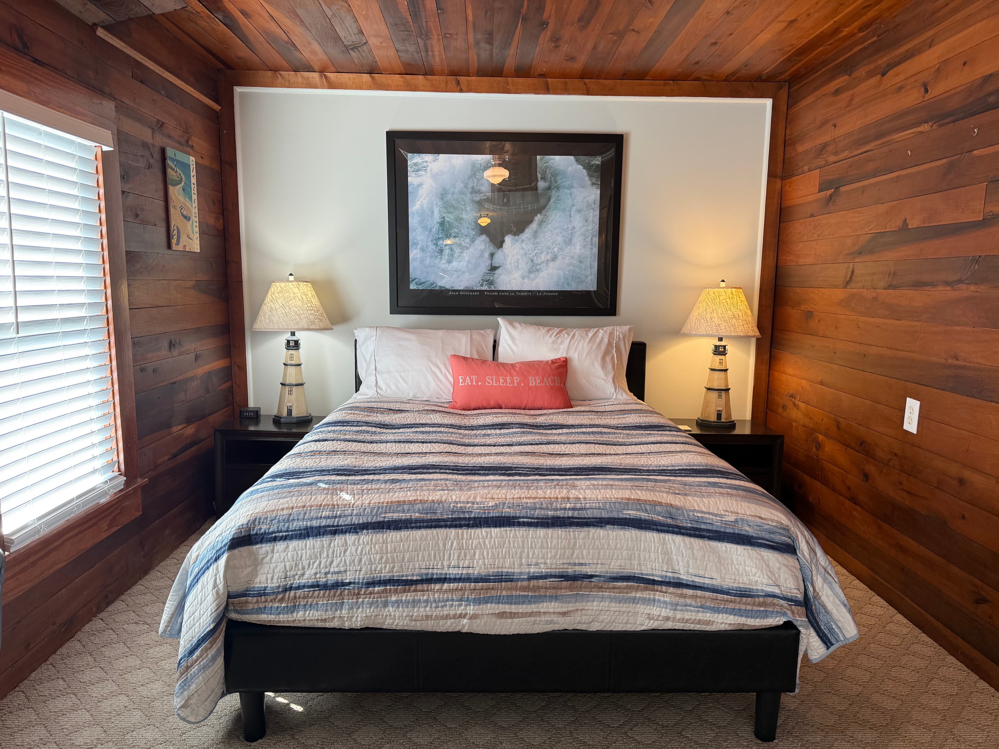 A rustic guest room featuring rich natural wood-paneled walls and ceiling. The bed is styled with a blue and white striped comforter and a coral accent pillow, flanked by two lighthouse-themed bedside lamps. A large framed picture hangs on the white accent wall above the headboard.