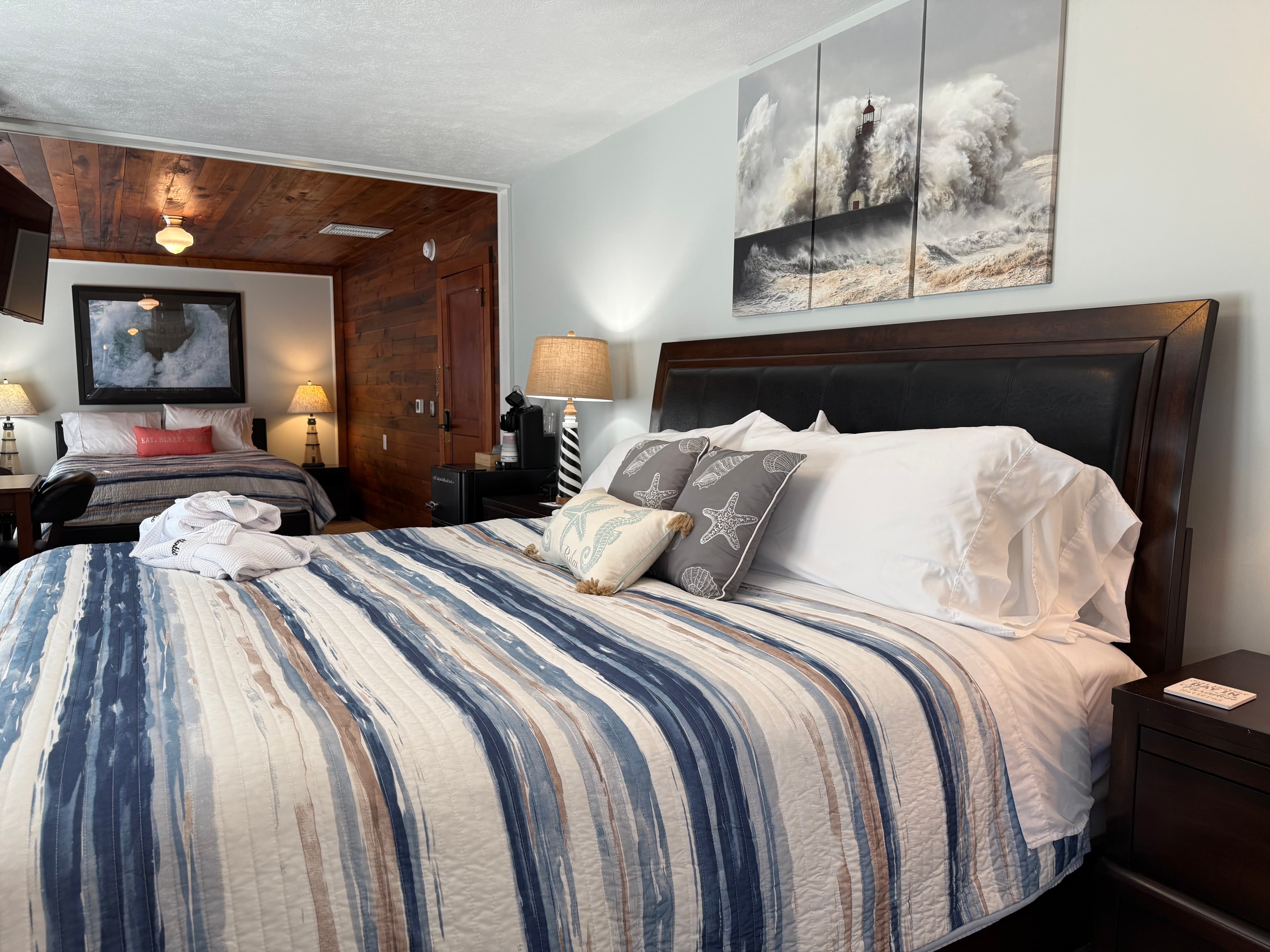 A suite featuring two beds with navy and white striped comforters. The foreground bed has a dark wood headboard and starfish accent pillows, while the background bed is situated in a cozy nook with warm wood-paneled walls and ceiling. A large three-panel canvas of a crashing ocean wave hangs above the main bed.