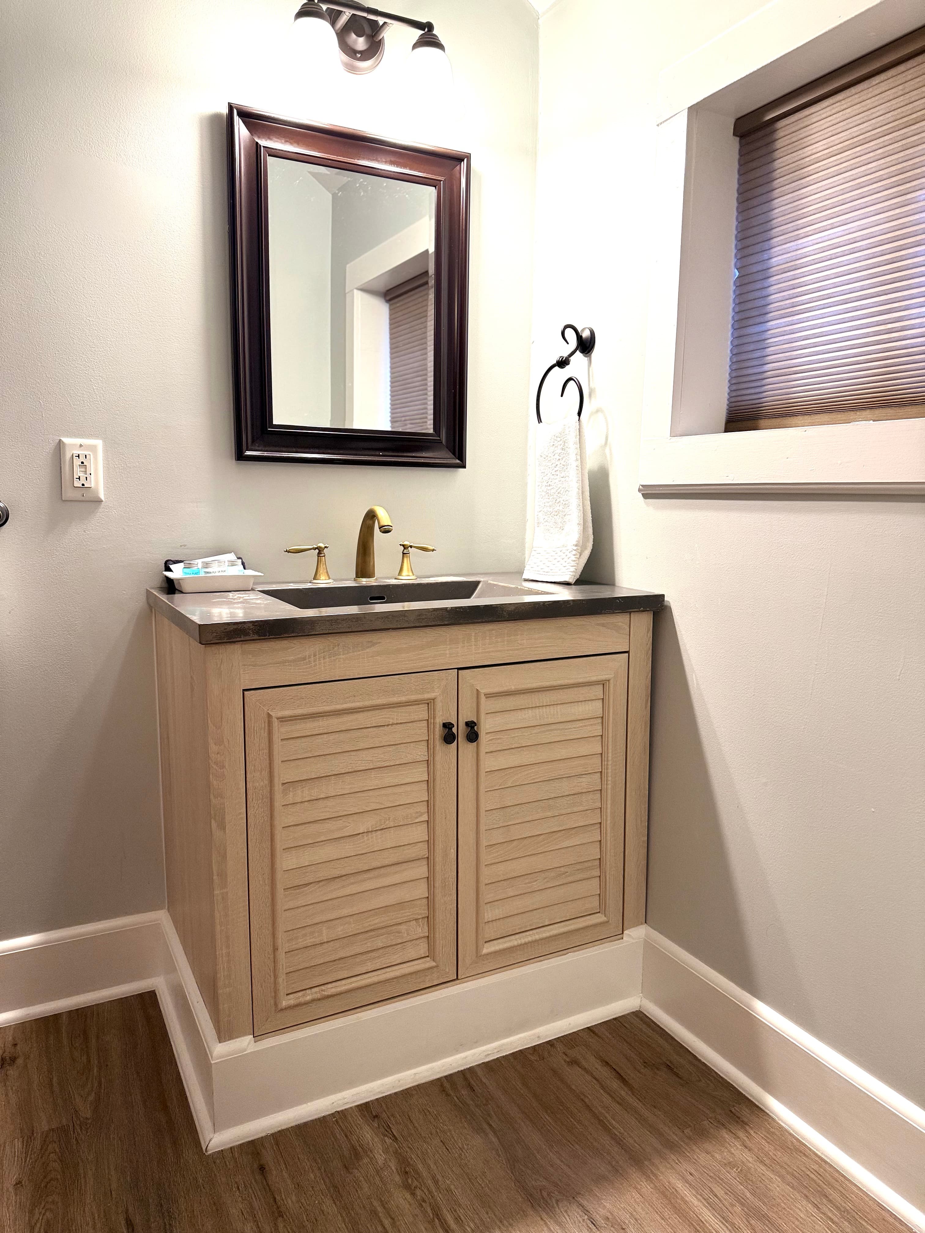 A modern bathroom vanity featuring light wood louvered cabinet doors and a dark countertop with a brass faucet. A dark-framed mirror hangs on the light gray wall above the vanity, and a window with a brown shade is visible to the right.