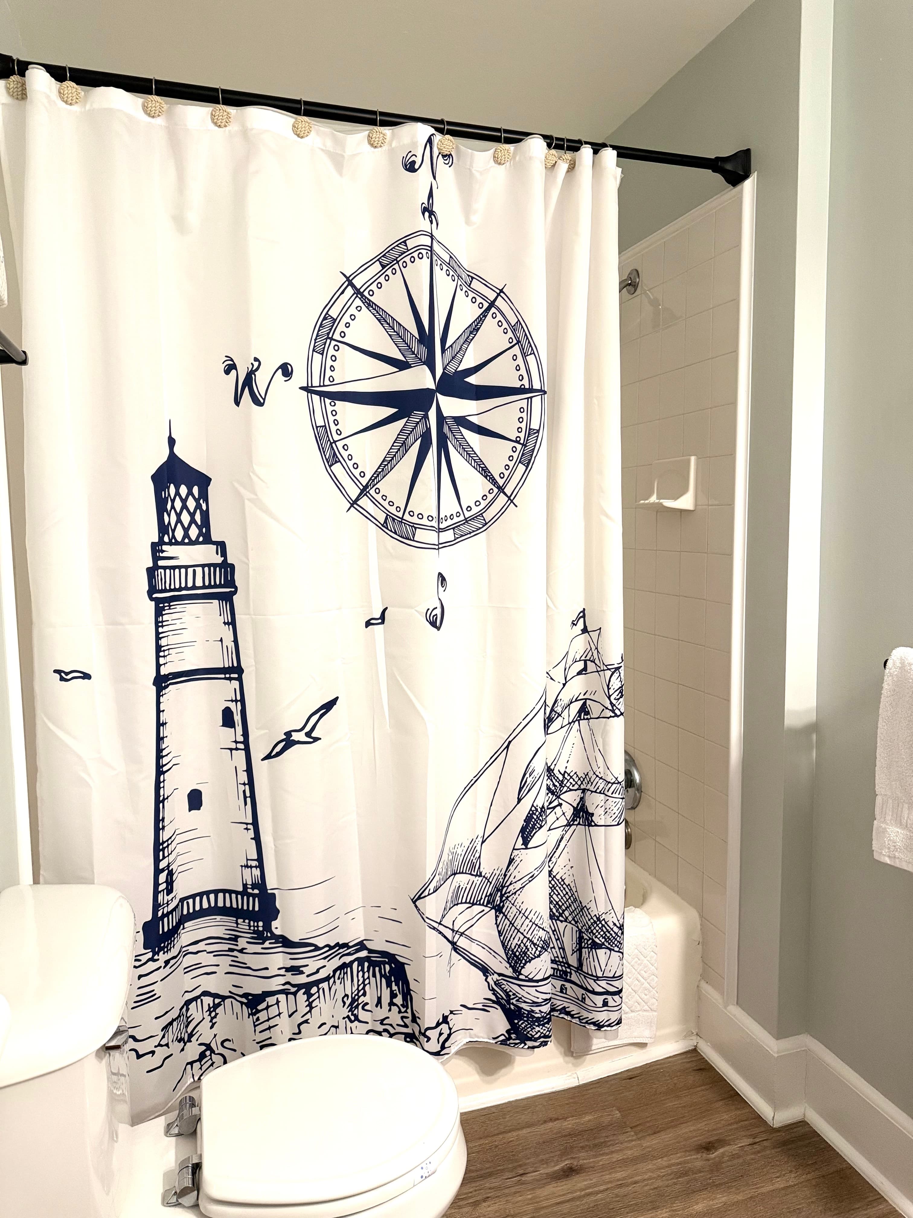 A white fabric shower curtain featuring a large navy blue nautical illustration of a lighthouse, a sailing ship, and a compass rose. The curtain is hanging in a bathroom with light gray walls and wood-style flooring.