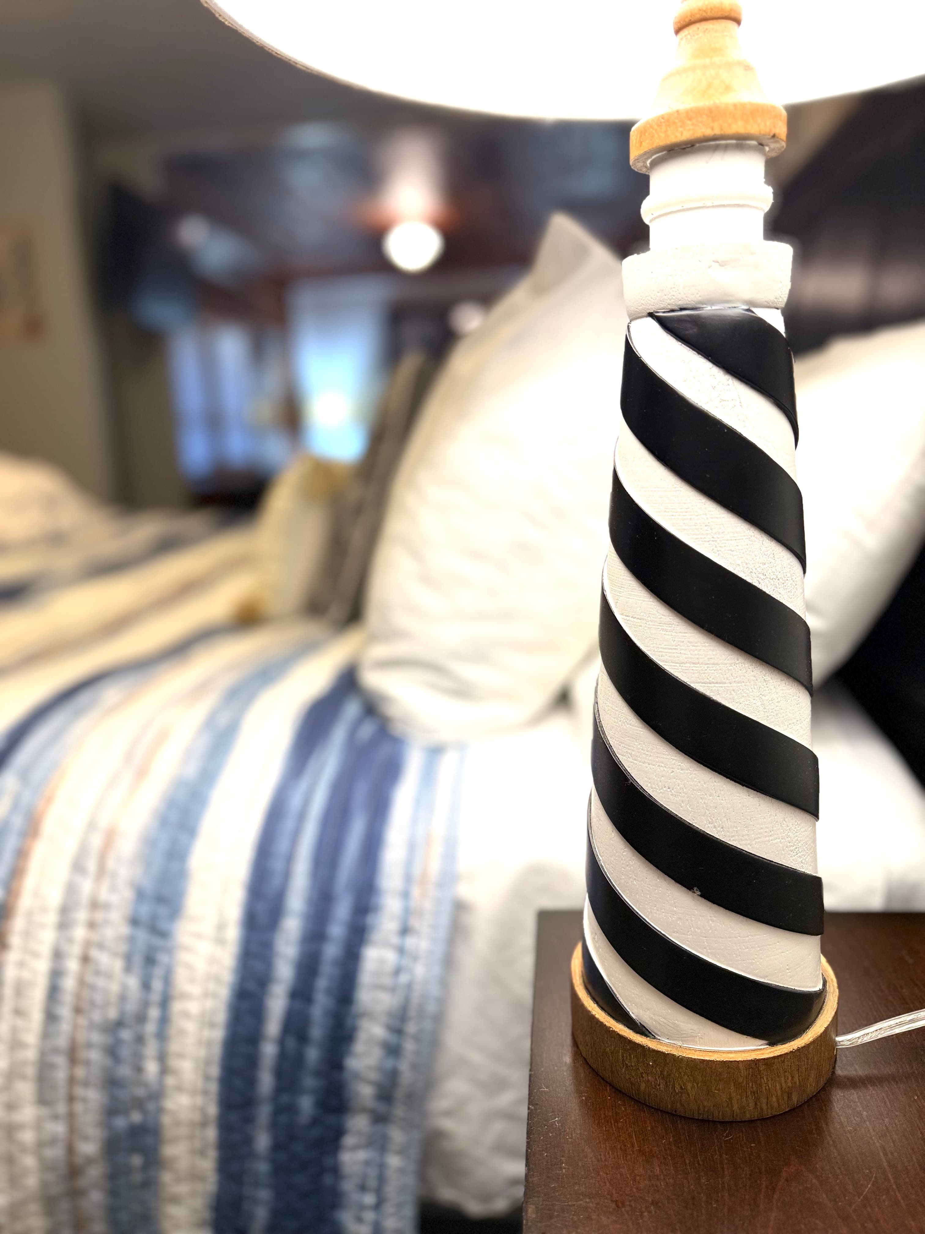 A bedside lamp in the shape of a lighthouse sits on a dark wood nightstand next to a bed with a blue and white striped comforter, with the soft glow of another room visible in the blurred background.