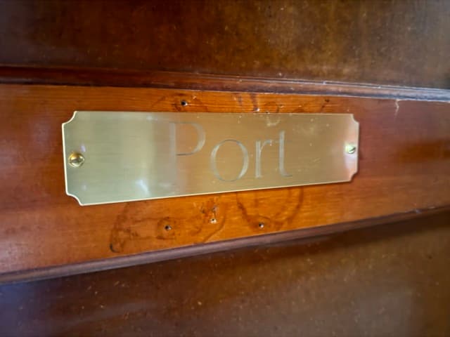 A close-up of a polished brass nameplate engraved with the word "Port" in a serif font, mounted on a rich, dark-toned wooden door.