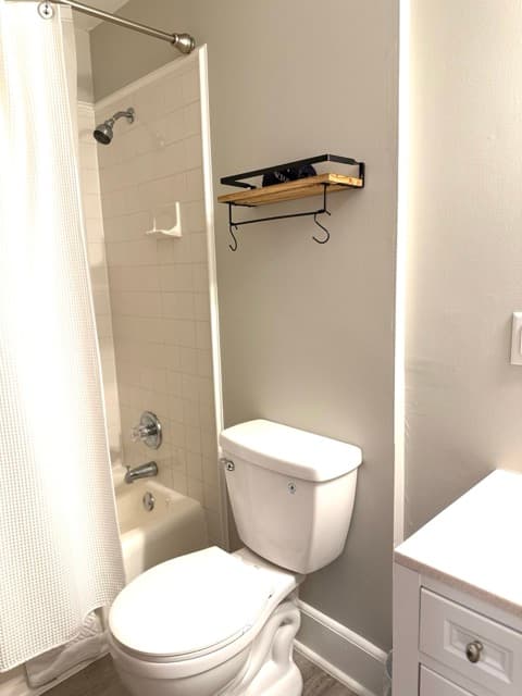 A clean, minimalist guest bathroom featuring a white toilet and a white bathtub with a matching shower curtain. A small wooden shelf with black metal hardware and hooks is mounted on the light gray wall above the toilet.