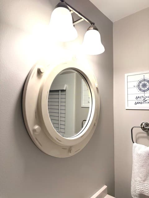 A close-up of a unique bathroom mirror, designed to look like a white circular ship's porthole. The mirror is mounted on a light gray wall beneath a triple-bulb vanity light, with a nautical-themed framed print visible in the background.