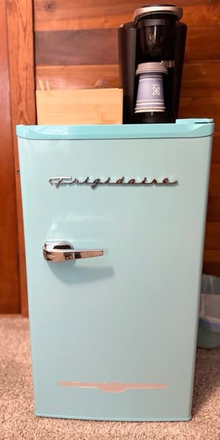 A close-up of a light blue retro-style Frigidaire mini-fridge featuring silver handles and trim. A black Keurig coffee maker with a white patterned cup sits on top, positioned against a rustic wood-paneled wall.