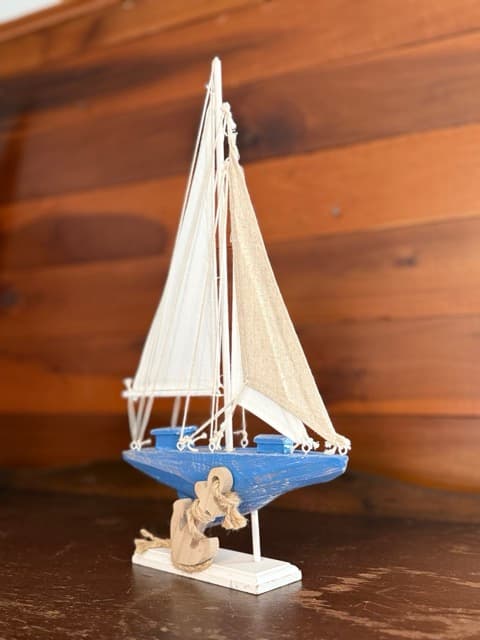 A close-up of a small decorative model sailboat with a blue hull, white sails, and tan rope accents. The model is displayed on a dark surface against a background of warm, natural wood-paneled walls.