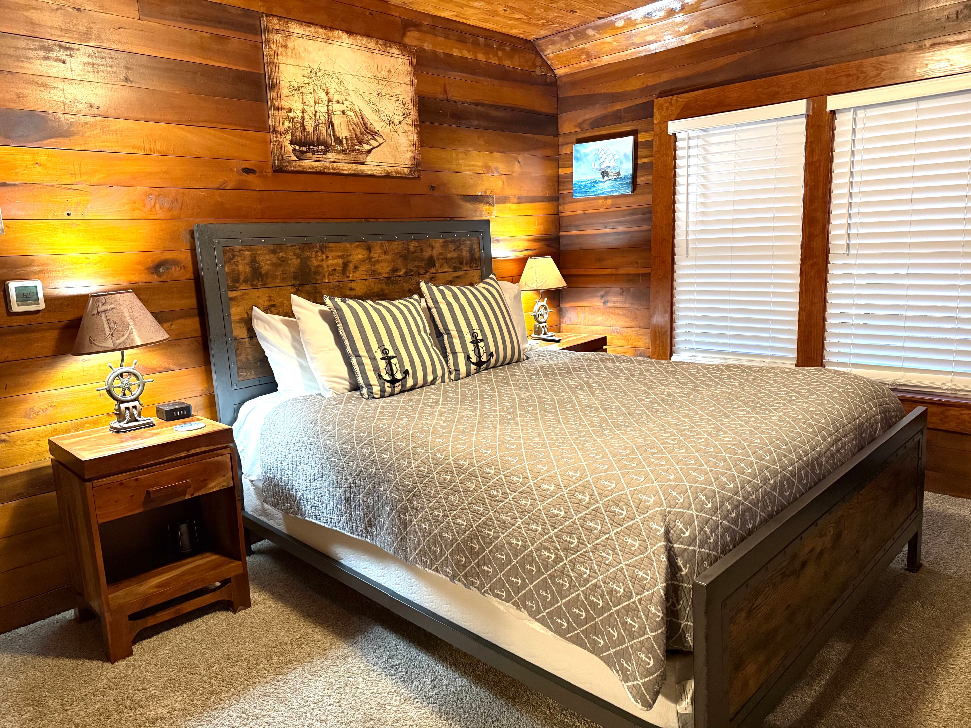 A rustic bedroom featuring horizontal natural wood-paneled walls and a queen bed with a dark wood frame and a gray patterned coverlet. The bed is accented with striped pillows featuring anchor motifs and is flanked by matching wood nightstands with nautical-themed lamps. A large vintage-style illustration of a sailing ship hangs on the wall above the headboard.
