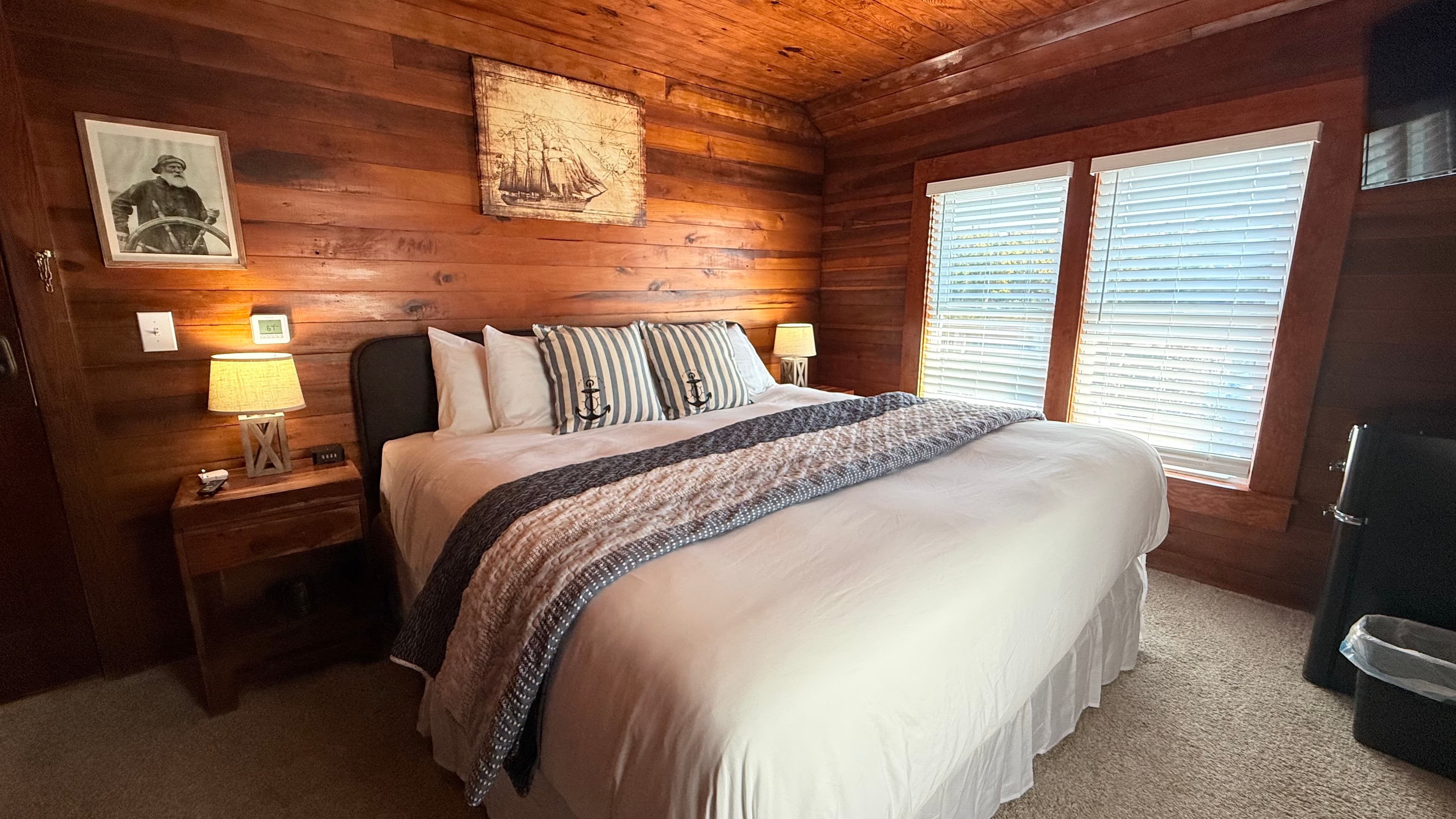 A rustic bedroom featuring horizontal natural wood-paneled walls and a queen bed with a dark wood frame and a gray patterned coverlet. The bed is accented with striped pillows featuring anchor motifs and is flanked by matching wood nightstands with nautical-themed lamps. A large vintage-style illustration of a sailing ship hangs on the wall above the headboard.