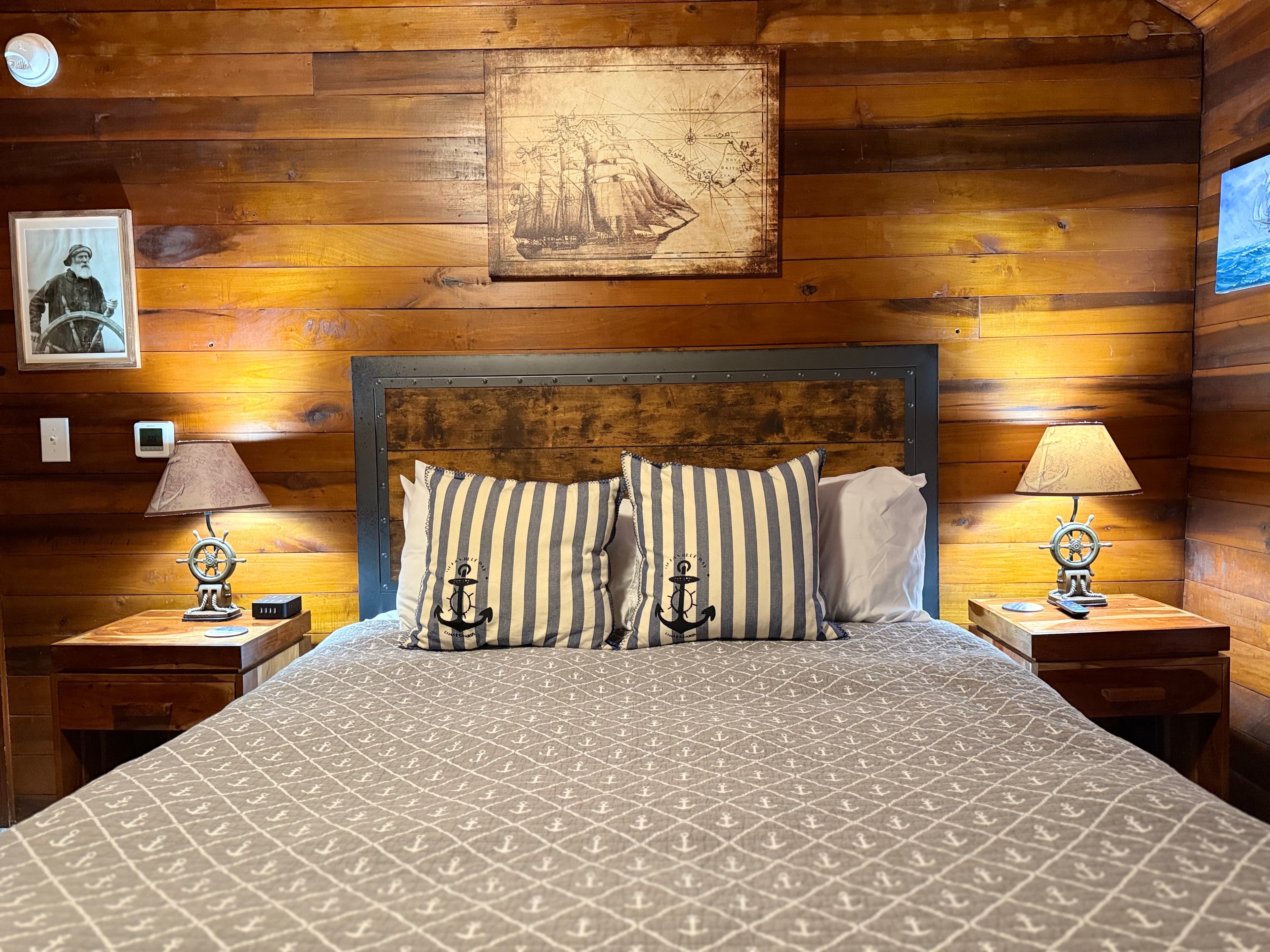 A guest bed featuring a large wooden headboard and a gray patterned coverlet, accented with striped pillows displaying anchor motifs. The room has rich natural wood-paneled walls and is decorated with vintage nautical art, including a large illustration of a sailing ship and a portrait of a sailor.