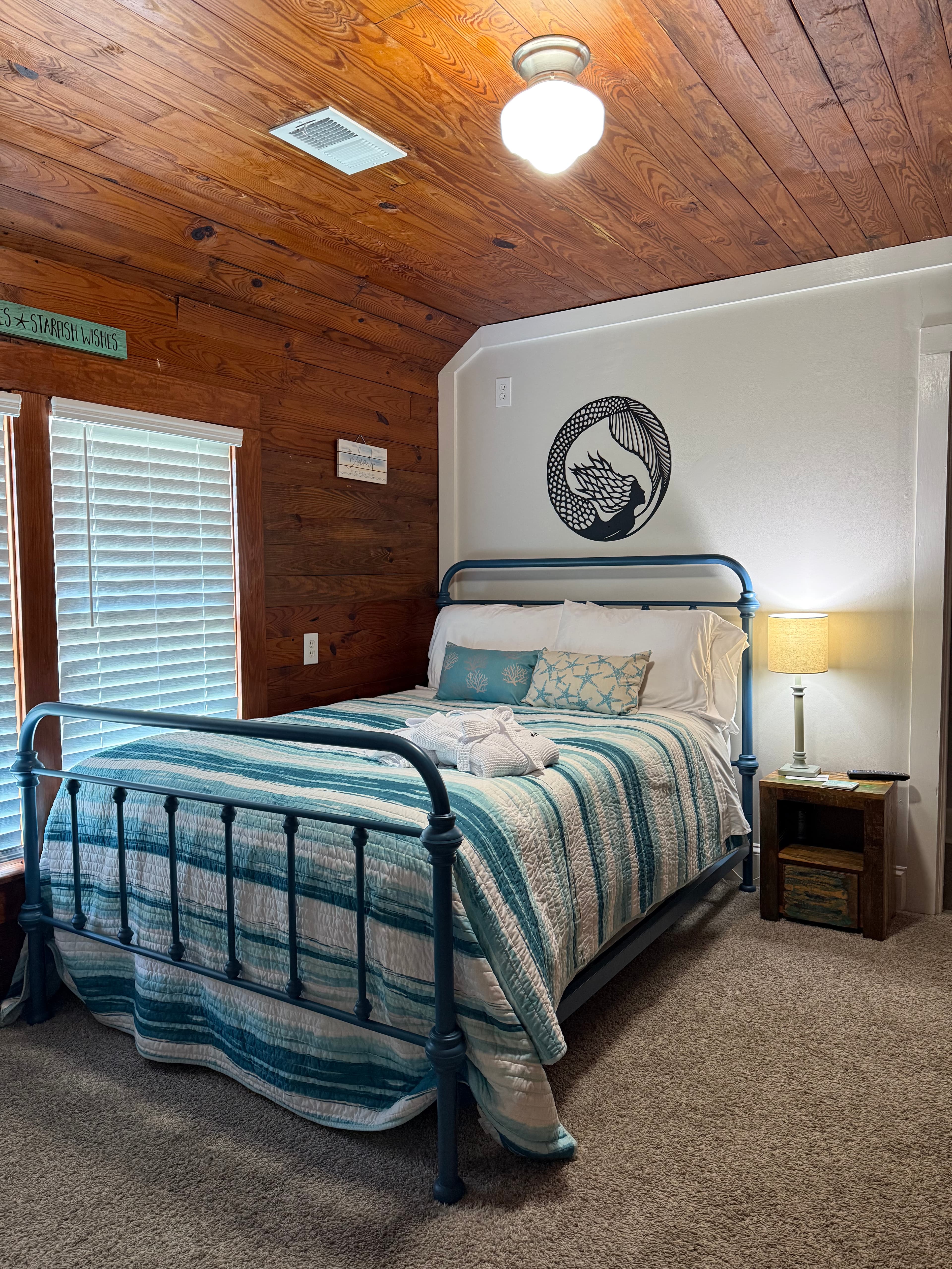 A nautical-themed guest room featuring a blue metal-frame bed with white bedding and a light blue accent pillow. One wall is covered in rich natural wood paneling, while the adjacent wall is a light gray and features a circular black-and-white illustration of a mermaid. The room is warmly lit by a ceiling globe light and a bedside lamp on a small wooden nightstand.
