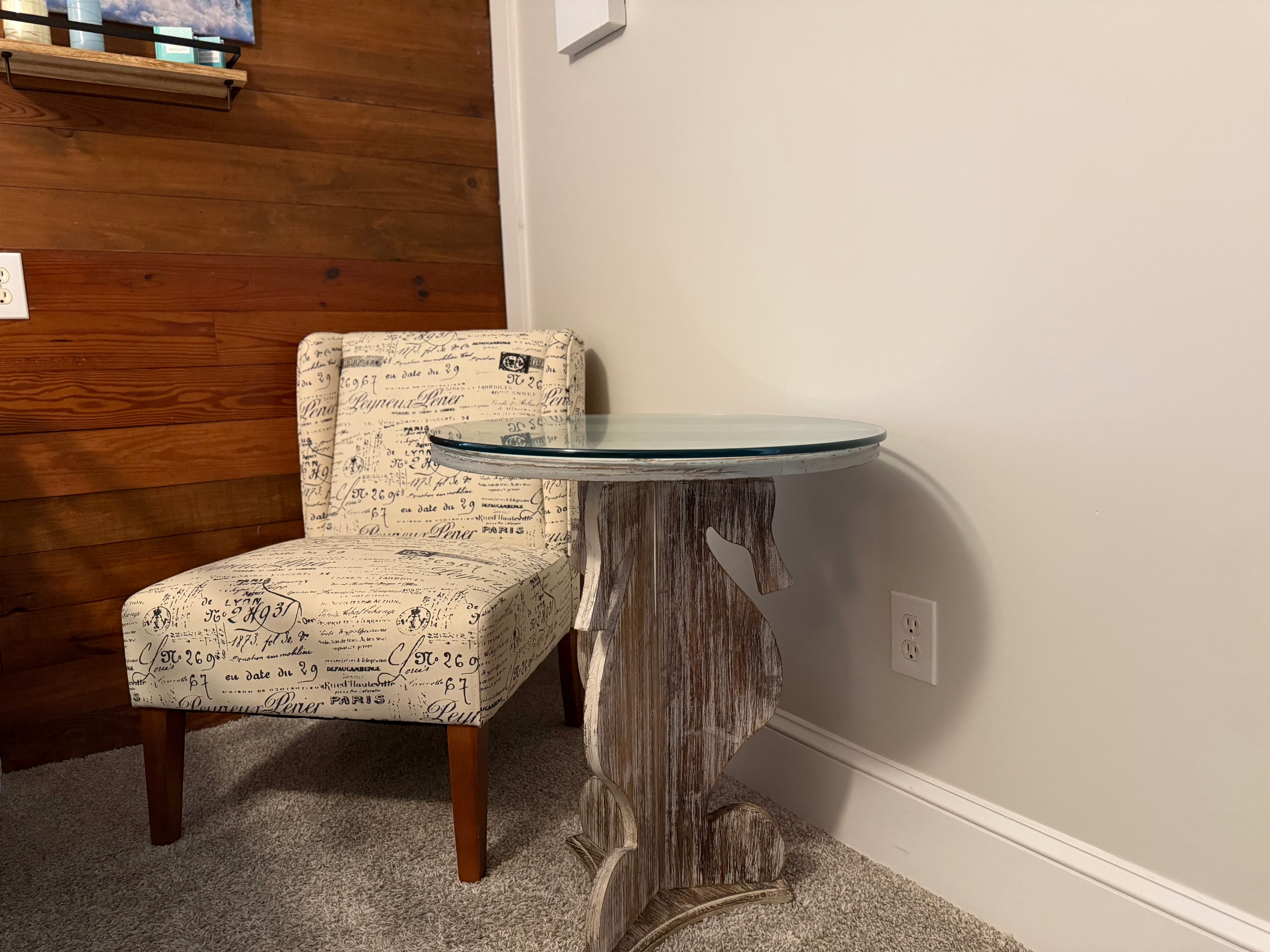 A cozy seating area in a guest room featuring a small armchair with vintage-style script upholstery and dark wood legs. Next to the chair is a round glass-top table with a driftwood-inspired base, set against a light gray wall and a natural wood-paneled accent wall.