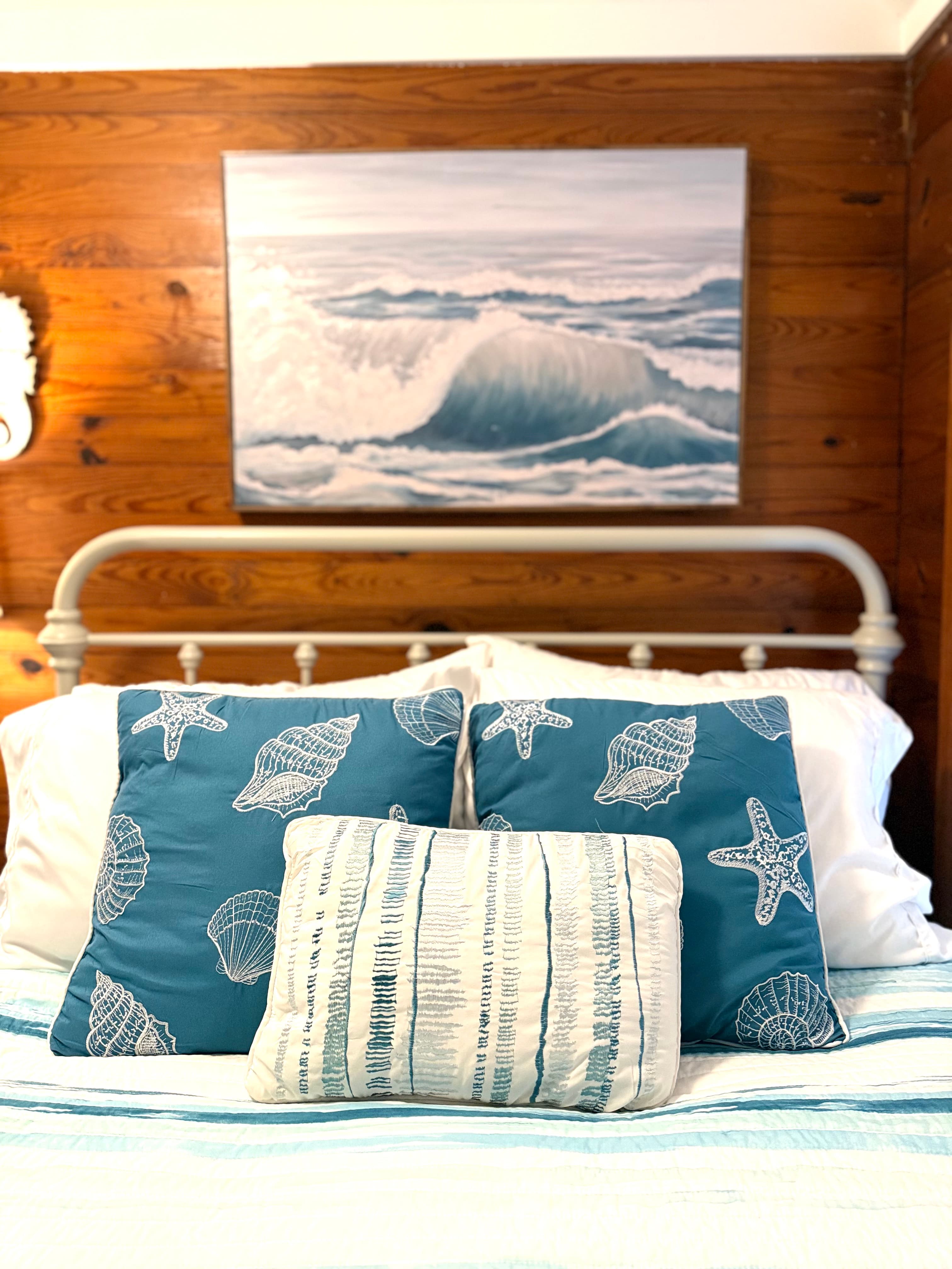 A close-up of a guest bed featuring a white metal headboard and a collection of nautical pillows, including two blue pillows with white seashell and starfish patterns. A large painting of a crashing ocean wave is mounted on the warm wood-paneled wall directly above the bed.