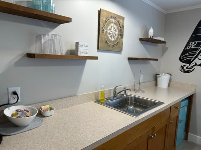 An updated kitchenette at The Atlantic Inn featuring a wooden wet bar with a stainless steel sink, floating wooden shelves with glassware, and a nautical-themed compass wall hanging.
