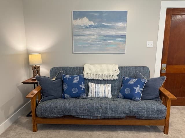 A cozy living area at The Atlantic Inn featuring a blue denim-style futon adorned with nautical seashell pillows, set against a neutral wall with a large coastal landscape painting and a classic wooden door.