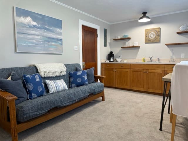 A brightly lit guest suite at The Atlantic Inn featuring a cozy living area with a blue denim-style futon and nautical pillows. The room includes a wooden kitchenette with floating shelves and a small dining table with white-covered chairs.