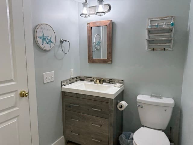 A modern bathroom at The Atlantic Inn featuring a gray wood vanity with a white vessel sink, a rustic wood-framed mirror, and nautical wall decor including a starfish print and tiered wire shelving.