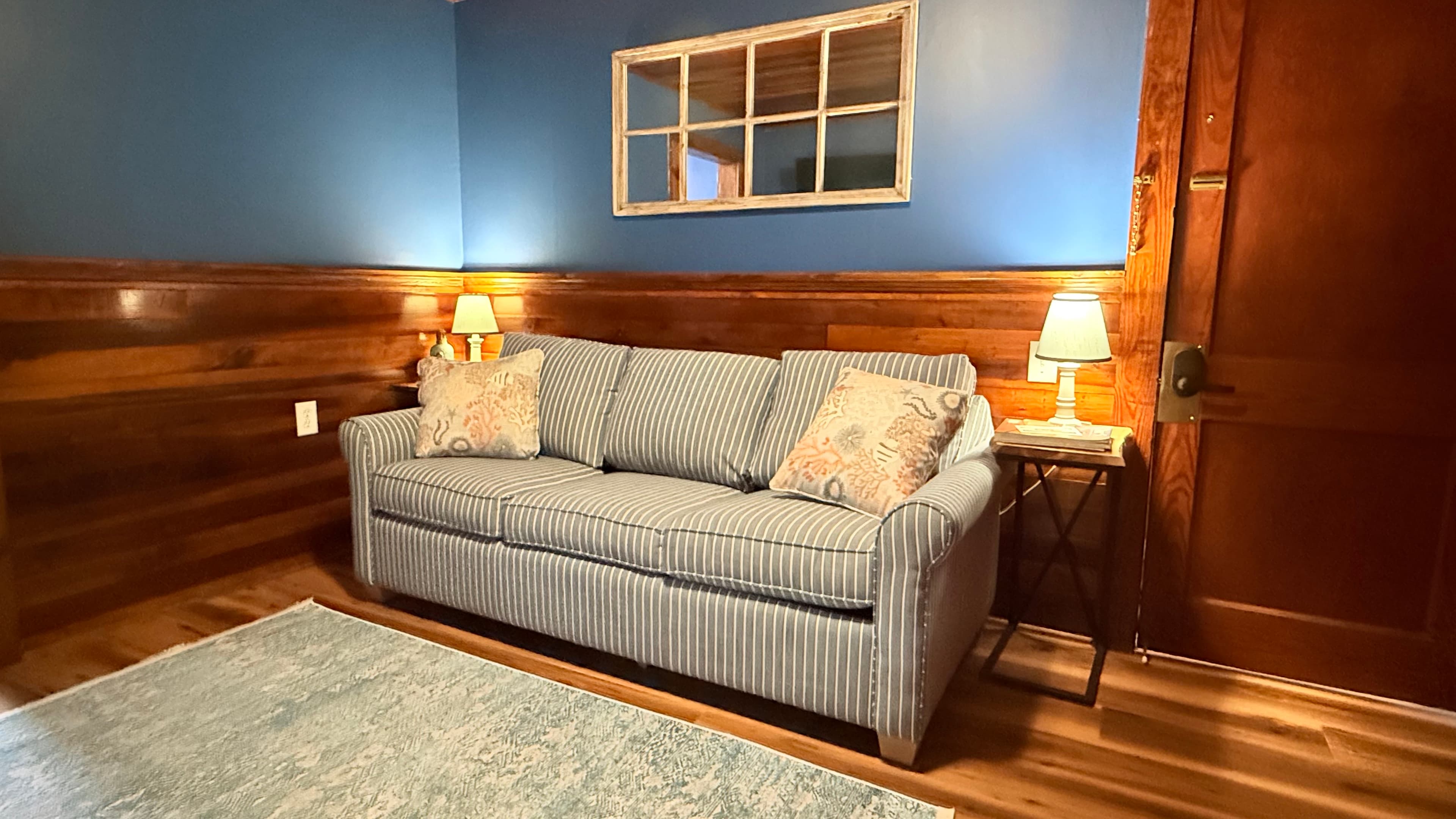 A rustic living area featuring wood wainscoting and a matching dark wood coffee table. The room includes a tan futon with nautical pillows, a striped wingback chair, and classic wooden doors.