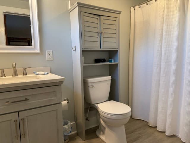 A modern bathroom featuring a light gray vanity with a white countertop and a storage cabinet over the toilet with louvered doors. A white shower curtain is visible to the right.