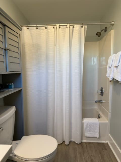 A bathroom featuring a white bathtub and shower combination with a simple white shower curtain. The room includes a toilet with a gray over-the-toilet storage cabinet and two white towels hanging on the adjacent wall.
