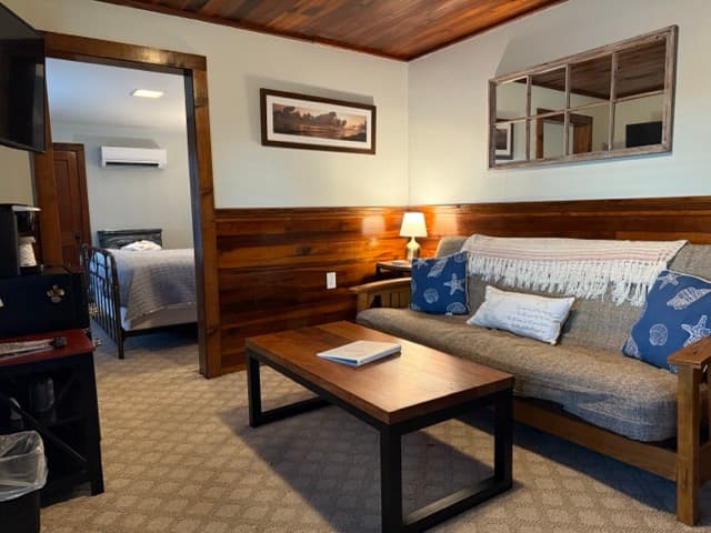 A rustic guest suite featuring original dark wood wainscoting and ceilings. The living area includes a tan futon with nautical pillows and a wooden coffee table, with a view into an adjoining bedroom with a metal bed frame.