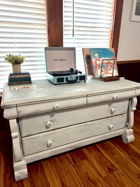 A light blue vintage-style suitcase record player sitting on a distressed white wooden dresser. Next to the player is a stand holding vinyl records, all positioned in front of a window with white blinds and dark wood trim.