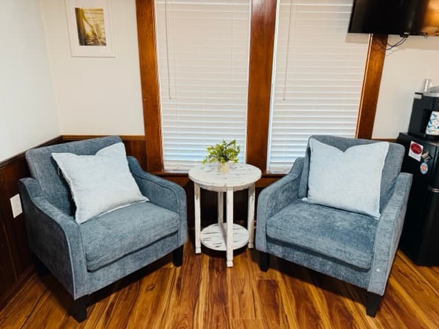 A cozy reading nook featuring two denim-blue upholstered armchairs with light blue pillows, centered around a small white round side table. The space is accented by warm wood flooring, wood-trimmed windows with white blinds, and a wall-mounted TV.
