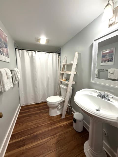 A brightly lit bathroom featuring a classic white pedestal sink, a white-framed mirror, and wood-style flooring. The room includes a white ladder-style shelf over the toilet, a white shower curtain, and fresh white towels hanging on the wall.