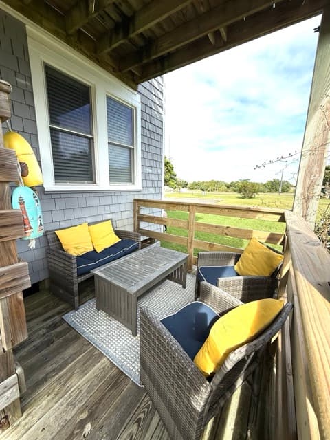 A private outdoor deck featuring gray wicker patio furniture with navy cushions and bright yellow pillows. The space includes a loveseat, two armchairs, and a matching coffee table on a gray rug, overlooking a grassy Hatteras landscape.