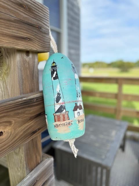 A close-up of a decorative light blue buoy hanging from a wooden post on a private deck. The buoy features hand-painted illustrations of local lighthouses.