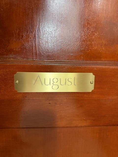 A close-up of a polished brass nameplate engraved with the word "August" in a serif font, mounted on a dark, rich-toned wooden door.