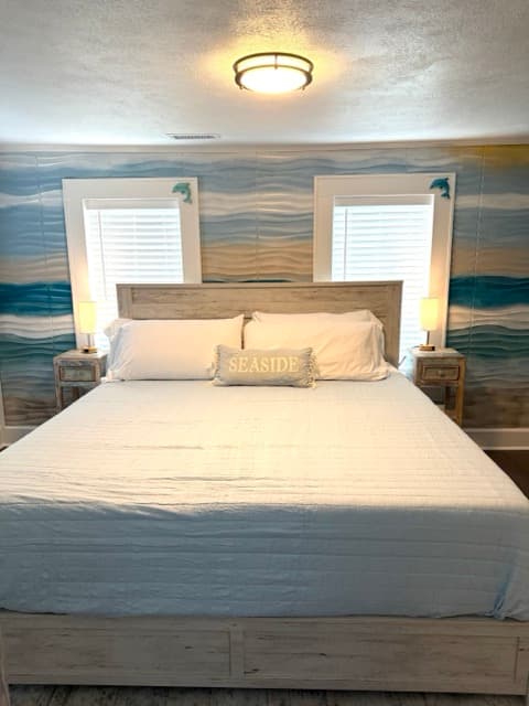 A brightly lit bedroom featuring a large bed with a light-colored wood headboard and white linens. The wall behind the bed is covered in coastal-themed wallpaper with blue, tan, and white horizontal waves, and two windows with white blinds flank the bed.