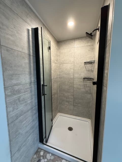 A modern walk-in shower featuring light gray wood-look tile walls, a white shower base, and a pebble-style floor. The shower is equipped with a black rainfall shower head and a glass door with a black frame.