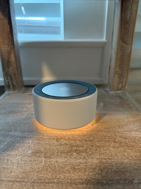 A close-up of a small, beige cylindrical noise machine sitting on a rustic wooden surface, casting a warm orange glow from its base.