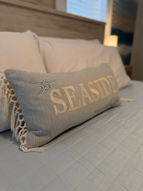 A close-up of a long, light blue decorative pillow on a bed, featuring the word "SEASIDE" embroidered in white and a small starfish detail. The pillow has white netted fringe on the ends and is set against a backdrop of crisp white bed linens and a rustic wooden headboard.