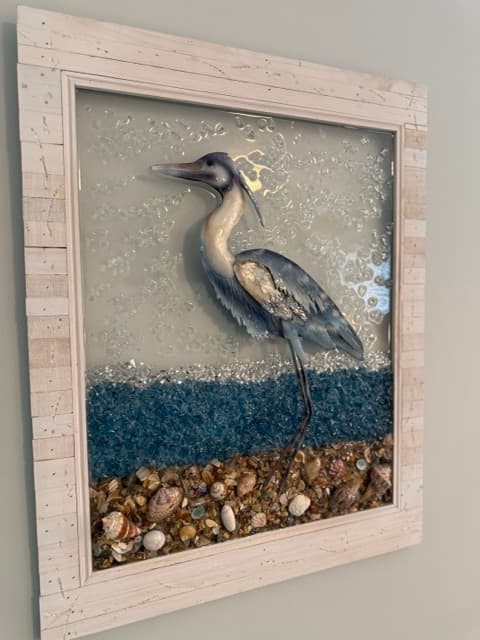 A piece of coastal 3D wall art at The Atlantic Inn featuring a blue heron standing in a textured scene made of sand, blue glass "water," and small seashells, all encased in a rustic white wooden frame.