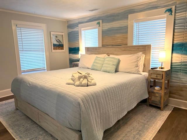 A brightly lit bedroom featuring a large bed with a light-colored wood headboard and white linens. The wall behind the bed is covered in coastal-themed wallpaper with blue, tan, and white horizontal waves, and two windows with white blinds flank the bed.