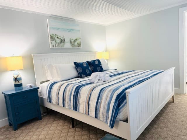 A guest room featuring a large white wooden bed frame with a navy and white striped comforter. The room includes a blue bedside table with a lamp, light gray walls, and a coastal landscape painting hanging above the headboard.