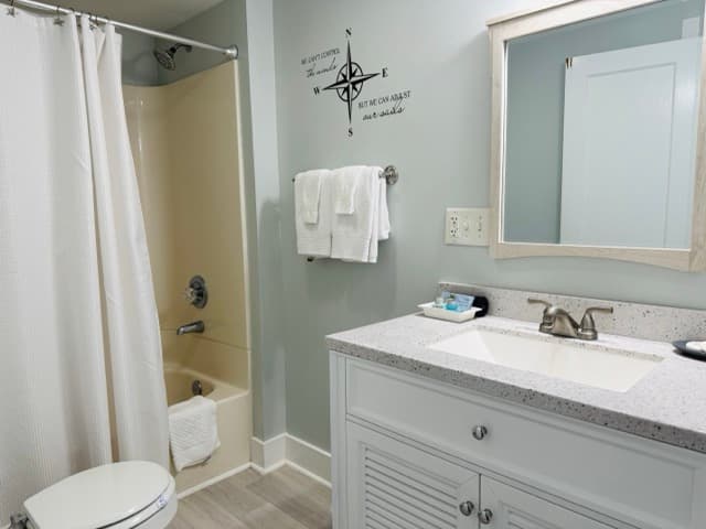A brightly lit bathroom featuring a white vanity with a louvered cabinet door and a white countertop. The light gray wall is decorated with a black compass decal and a quote, and the room includes a bathtub with a white shower curtain.