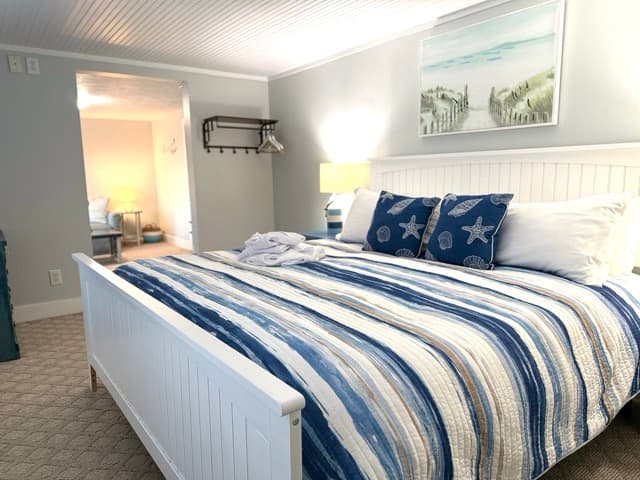 A brightly lit bedroom featuring a large bed with a white wooden headboard and a navy and white striped comforter. The room includes blue starfish accent pillows, light gray walls, and a coastal painting of sand dunes hanging above the bed.