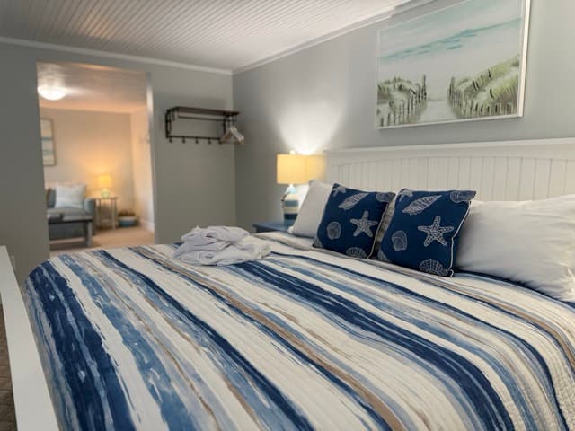 A brightly lit bedroom featuring a large bed with a white wooden headboard and a navy and white striped comforter. The room includes blue starfish accent pillows, light gray walls, and a coastal painting of sand dunes hanging above the bed.