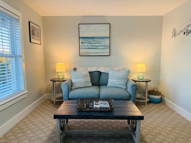 A cozy seating area featuring a light blue loveseat with striped accent pillows and a dark wood coffee table. The room is decorated with a coastal painting on a light gray wall, two bedside-style lamps on round side tables, and a window with white blinds.