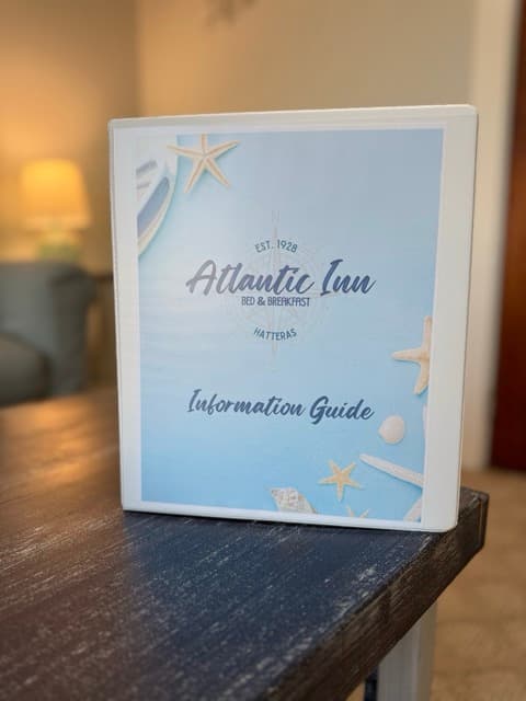 A close-up of a white three-ring binder sitting on a dark wood table. The cover of the binder is light blue with starfish graphics and features the text "The Atlantic Inn Information Guide."