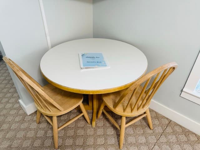 A small circular white dining table tucked into a corner at The Atlantic Inn, paired with two light wood spindle-back chairs. The "Atlantic Inn Information Guide" binder sits in the center of the table.