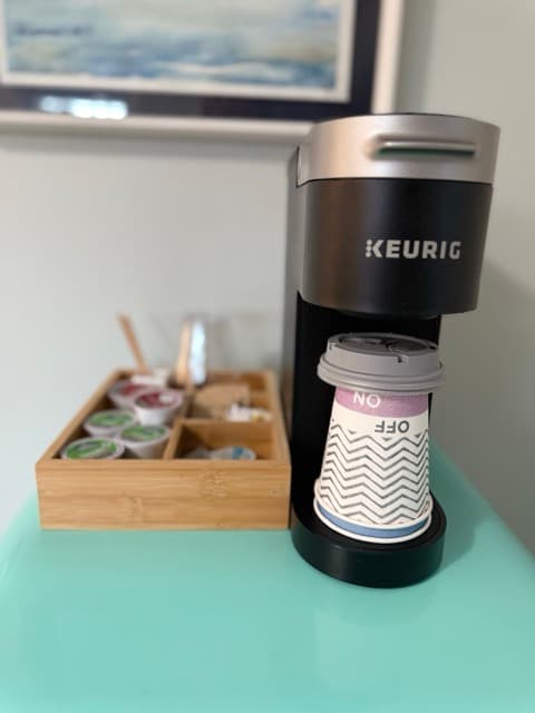 A close-up of a black Keurig coffee maker sitting on a turquoise surface. A patterned paper cup is placed in the machine, and a wooden organizer containing various coffee pods and stir sticks sits to the left.
