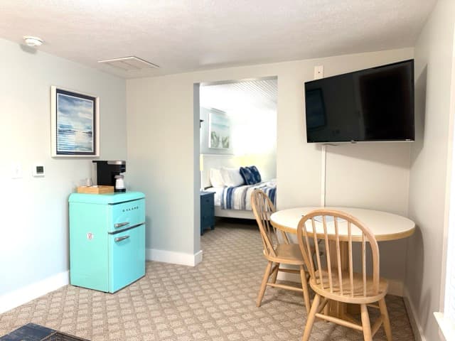 A suite featuring a dining area with a small white circular table and two light wood chairs. The room includes a turquoise mini-fridge with a Keurig coffee maker on top, a wall-mounted TV, and a view into an adjacent bedroom with a striped navy and white comforter.