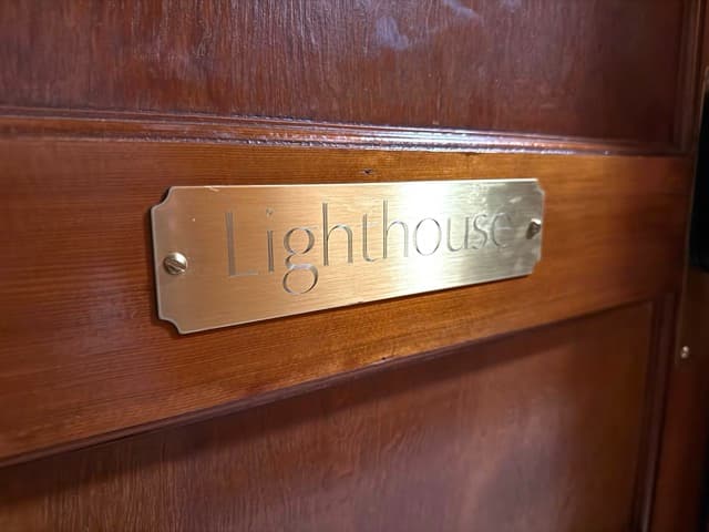 A close-up of a polished brass nameplate engraved with the word "Lighthouse" in a serif font, mounted on a dark, rich-toned wooden door.