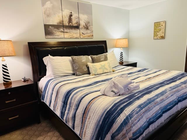 A large guest bed featuring a dark wood headboard, a navy and white striped comforter, and nautical-themed accent pillows. The room includes two black-and-white striped lighthouse-style lamps on dark wood nightstands and a three-panel canvas of a crashing ocean wave hanging above the bed.